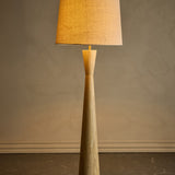 Leonor Wooden Floor Lamp with Linen Shade in Natural, showing the tapered rubberwood column with pinched waist and tan linen drum shade, set against a plain wall.