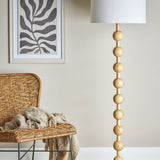 Giacoma Wooden Floor Lamp in natural wood with stacked sphere stem and white linen drum shade, shown beside a rattan chair in a living room.
