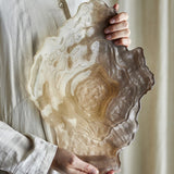 Aiku Shell Glass Bowl in pearl, a wide shallow dish with rippled, shell-like rim and ridged centre, held in two hands against a neutral linen backdrop.