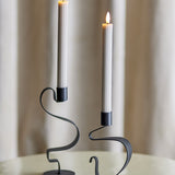 Ponza LED Candles in beige, two tall ribbed paraffin-wax columns with flame-shaped tips, displayed lit in black S-curve candle holders against a curtained backdrop.