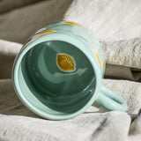 Green Agnes Stoneware Cup 230ml tipped on woven fabric, showing glossy interior with hand-painted lemon motif and matching exterior lemon pattern.