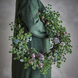 Cedar Artificial Christmas Wreath, 50cm, with frosted green sprigs, pinecones, and berry clusters, draped over a model's arm in a green coat.