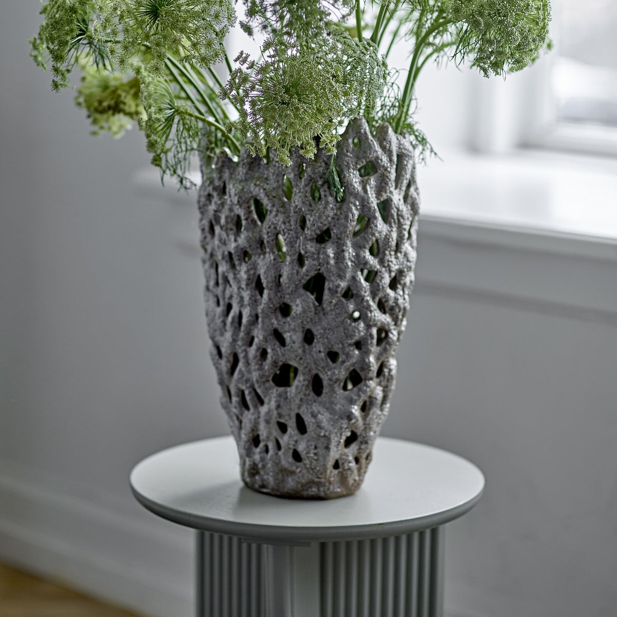 Eunice Stoneware Vase 37cm in White with pierced, textured coral-like walls, holding greenery on a grey pedestal table in a softly lit room.