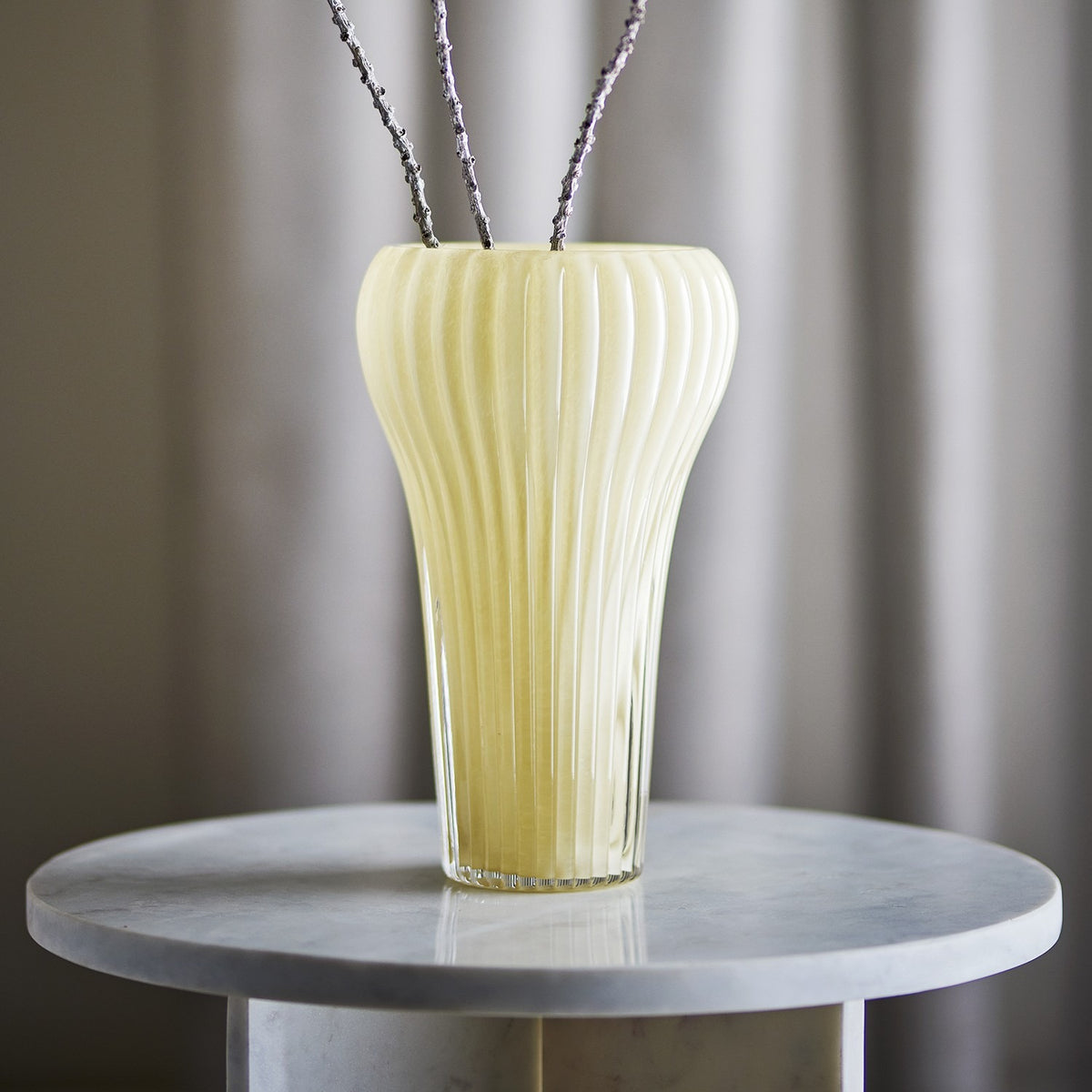 Averie Glass Vase 28cm in yellow, mouth-blown with fine vertical ribs and a curved silhouette, shown on a marble side table with dried branches.