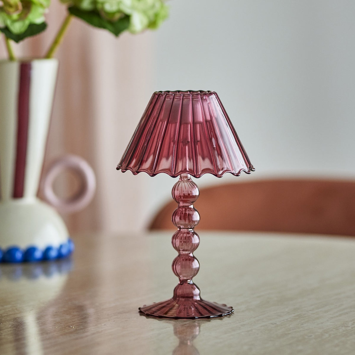 Pink glass tealight holder shaped like a mini lamp with a fluted wavy shade, stacked bead stem, and ribbed scalloped base, on a wooden table near a vase of flowers.