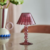 Pink glass tealight holder shaped like a mini lamp with a fluted wavy shade, stacked bead stem, and ribbed scalloped base, on a wooden table near a vase of flowers.