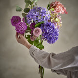 Edge Artificial Bouquet in Purple held by hand, showing hydrangea-style clusters, pompom heads and eucalyptus foliage against a plain grey background.