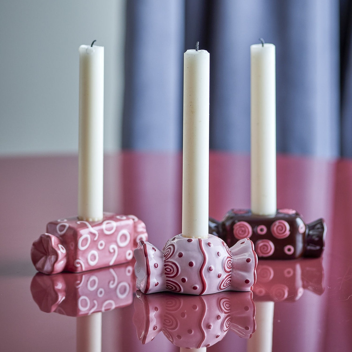 Maiano Wrapped Sweet ceramic candle holders, set of 3 in pink and dark plum with embossed swirls, rings and dots, each holding a slim white taper candle on a glossy pink surface.