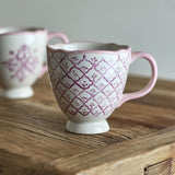 Stoneware mug from the Wonderland Christmas set, footed base, scalloped rim and curved handle in pink, decorated with a purple trellis pattern, shown on a wooden surface.