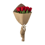 Artificial tulip bouquet in red with cupped heads on long stems and broad green leaves, wrapped in kraft paper and tied with ribbon, on a plain white background.
