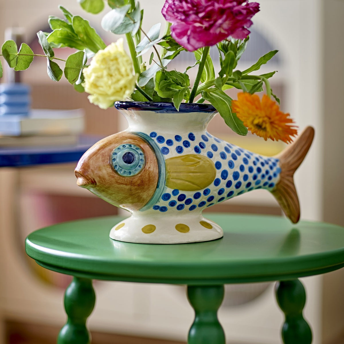 Nobu Fish Stoneware Vase in multicoloured ceramic with hand-painted dot scales, round eye and ridged tail, filled with fresh flowers on a green side table.