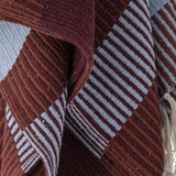 Isnel Red Striped Throw in multicoloured woven stripes, showing a ribbed fabric texture with diagonal pinstripes in burgundy, rust and pale blue, draped in folds with a knotted yarn fringe visible.