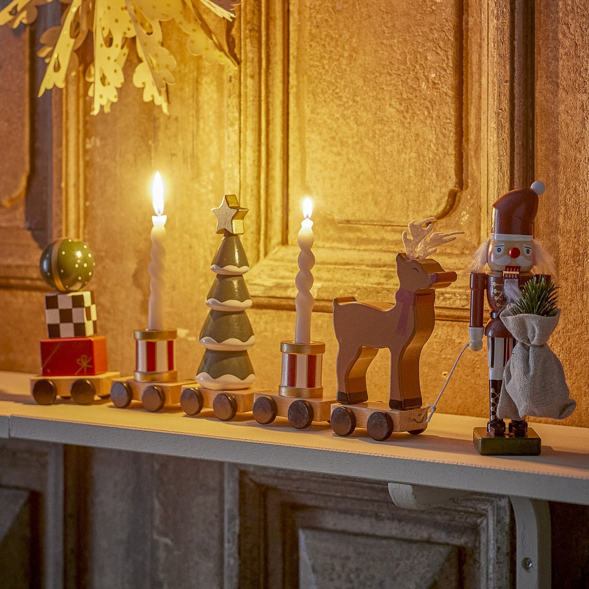Vadim Christmas Decoration wooden train on a mantel, with bauble-topped gift wagon, striped candle wagons, snowy tree, reindeer and nutcracker figure, displayed against a golden panelled wall.