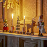 Vadim Christmas Decoration wooden train on a mantel, with bauble-topped gift wagon, striped candle wagons, snowy tree, reindeer and nutcracker figure, displayed against a golden panelled wall.