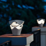 Cornet B/27 Cone LED bollard light in Stone Grey, tiered wavy layers glowing warm white, shown outdoors on a stone ledge at dusk.