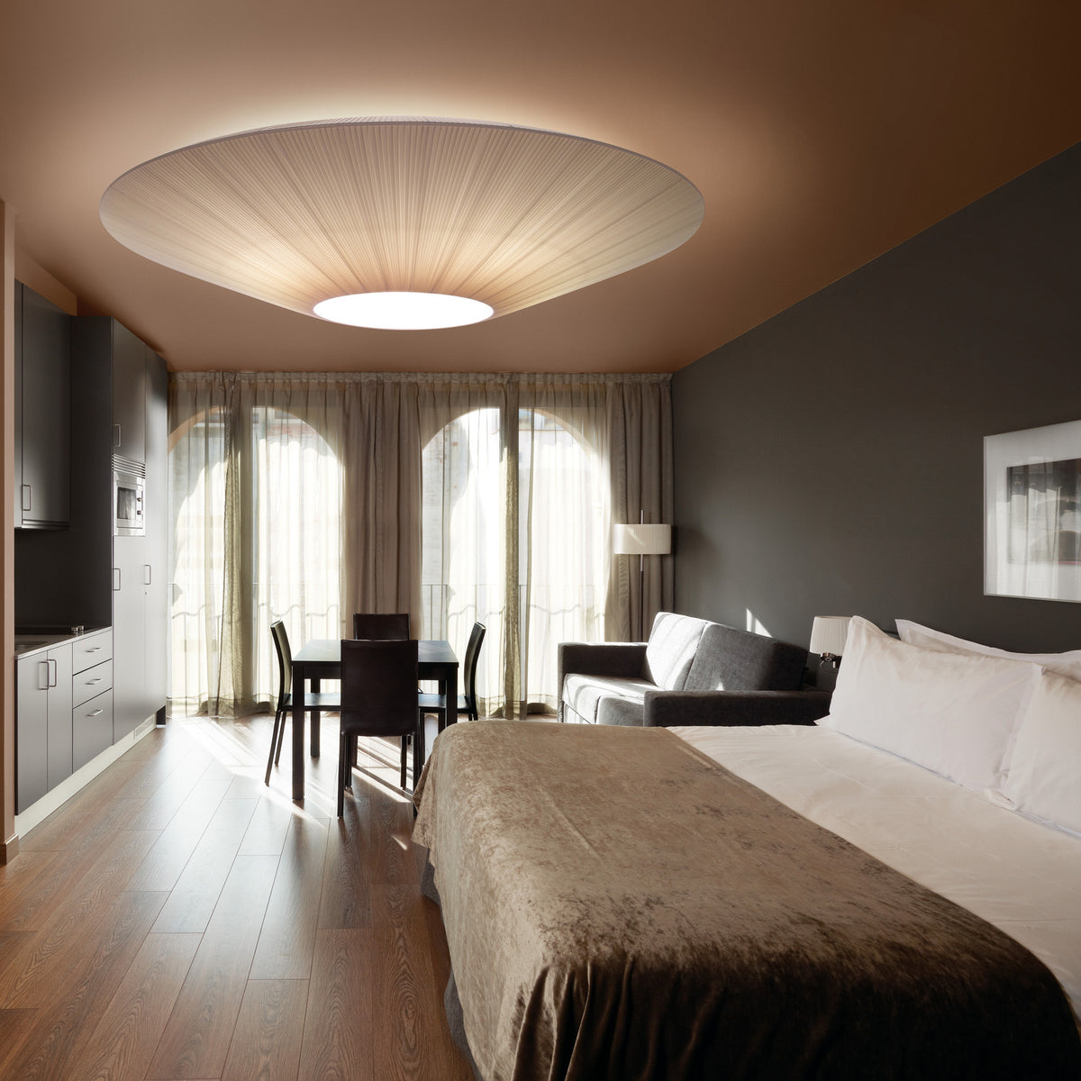 Siam 80 Ribbon Cone Ceiling Light in cream, a wide pleated semi-flush fixture glowing above a bedroom with grey walls and a brown velvet bed throw.