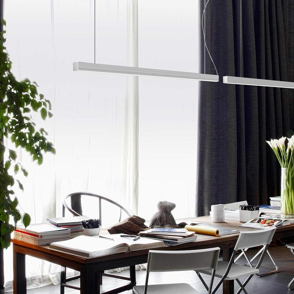 Brooklyn Line Aluminium Linear LED Pendant Light, 152cm slim bar in brushed metal finish, hung above a wooden dining table in a living space with dark curtains and plants.