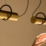 Solid SH Sens Cylinder Pendant Light in brass, slim cylindrical tube suspended on thin wires with a black ring bracket, shown against a warm neutral wall.