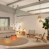 Nans Bag/38 Wood LED outdoor floor lamp with woven brown lattice shade and teak handles, shown glowing on a rug in a modern living room with pendant lights and lounge chairs.