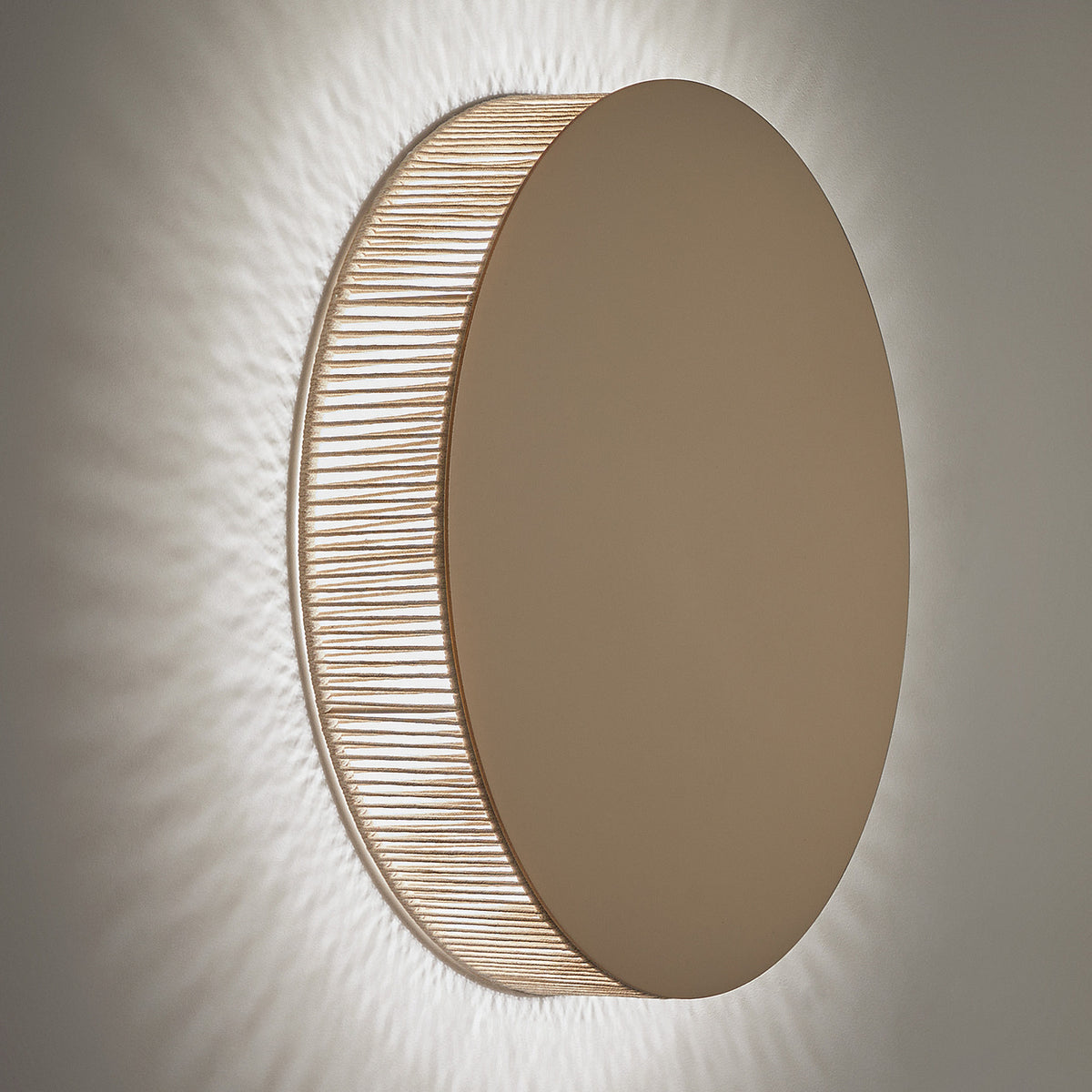 Helios A/02 round wall light with flat oak brown disc face and beige cotton thread edge, integrated warm white LED casting a soft glow on a plain grey wall.