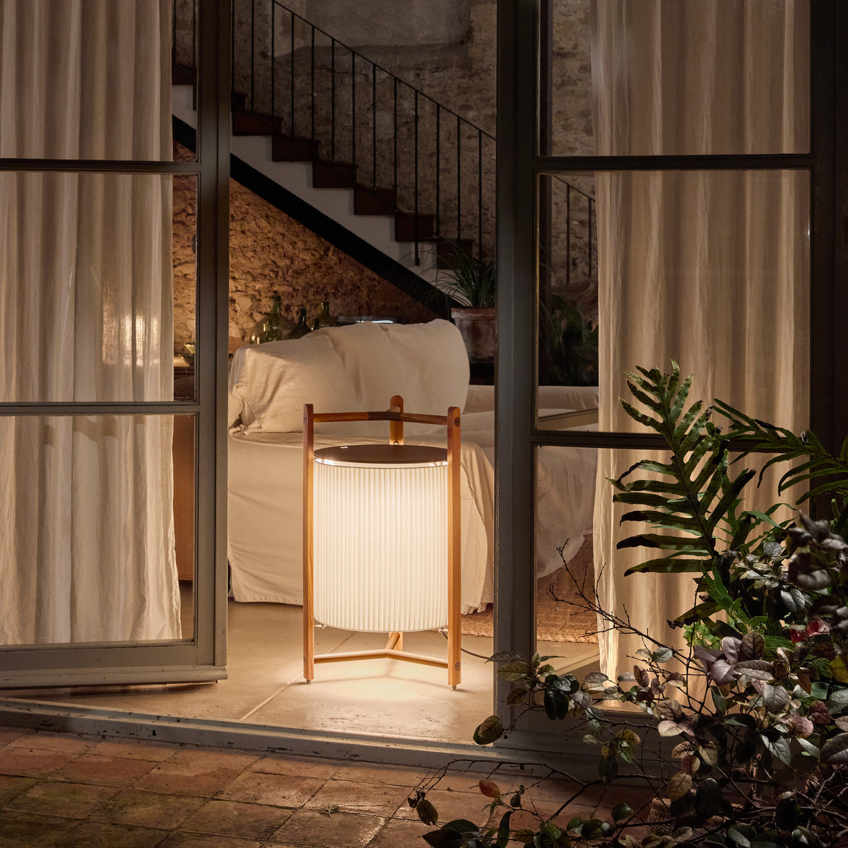 Kando P/76 Ribbon LED Floor Lamp in white, pleated cylindrical shade within an open teak wood frame, glowing beside a sofa in a living room at night.