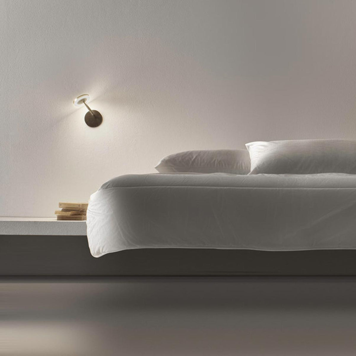 Bella Direct LED wall light with round disc head and angled arm in bronze finish, glowing softly above a bedside in a minimalist bedroom setting.
