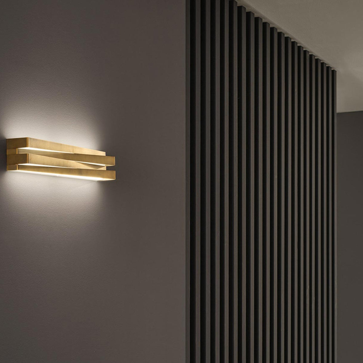 Cross Glass LED Wall Light 30cm in brushed brass finish, stacked crossing bars mounted on a grey wall, glowing warm white light beside a dark slatted wood panel.