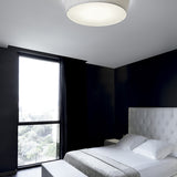 Plafonet 60 Ribbon LED ceiling light with pleated white drum shade mounted flush on a bedroom ceiling above a white tufted headboard and dark walls.