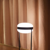 Beaded T Glass LED Table Lamp with an opal white, flattened round shade held by a dark central band between two slim black rods, shown lit against a pink curtain backdrop.