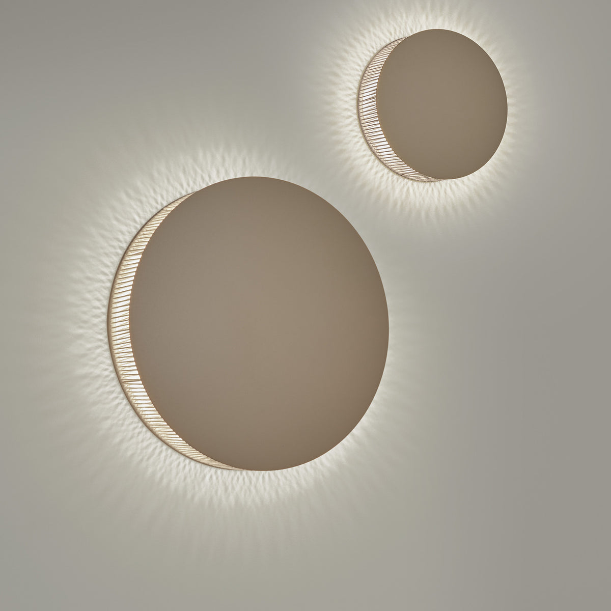 Helios A/01 round LED wall lights in Oak Brown & Beige, two sizes shown with brown disc fronts and beige cotton thread edges casting a soft radial halo on a light grey wall.