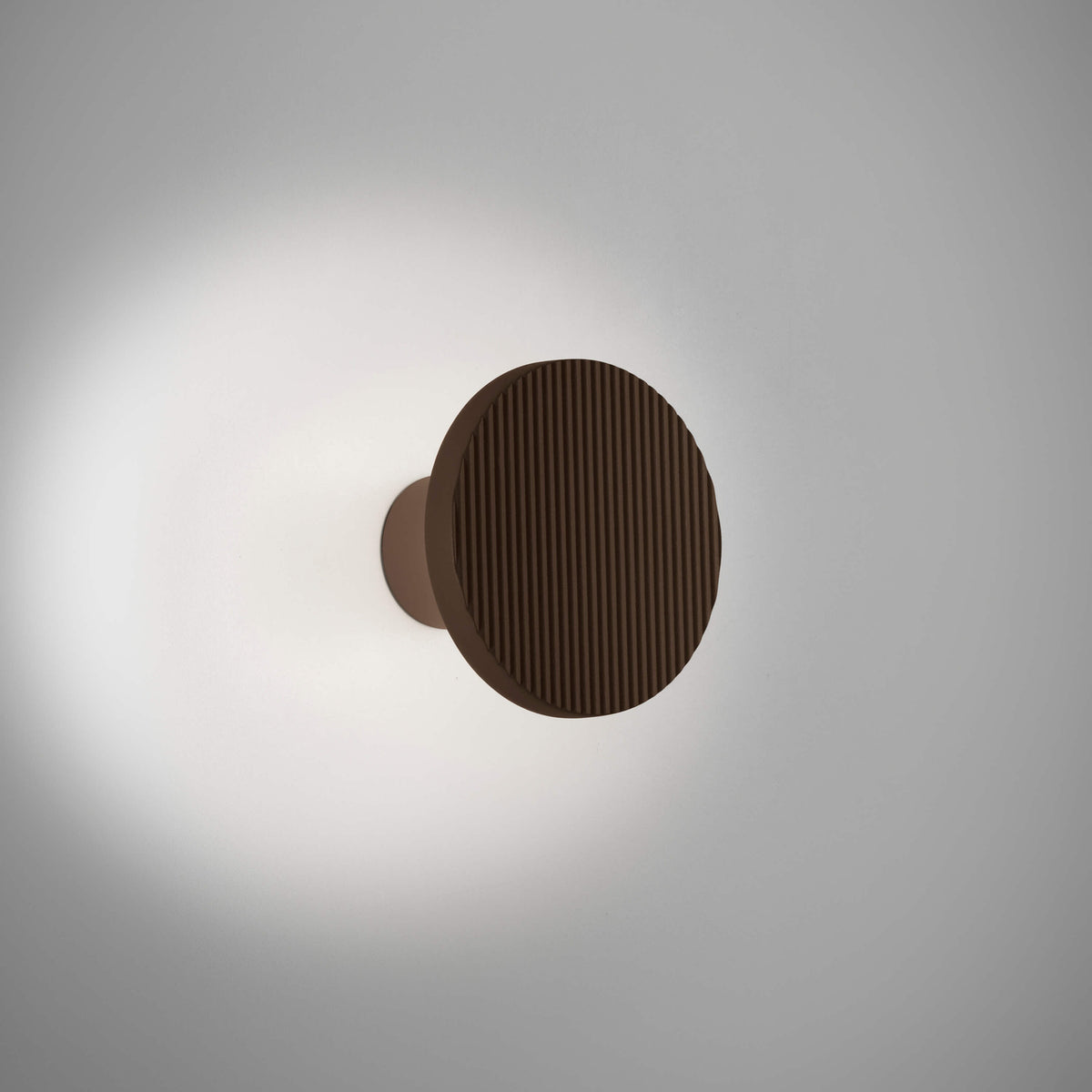 Arid A/01 round aluminium LED wall light in Textured Graphite Brown, fluted disc on a short stem casting a soft glow on a plain grey background.