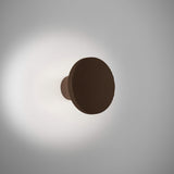 Arid A/01 round aluminium LED wall light in Textured Graphite Brown, fluted disc on a short stem casting a soft glow on a plain grey background.