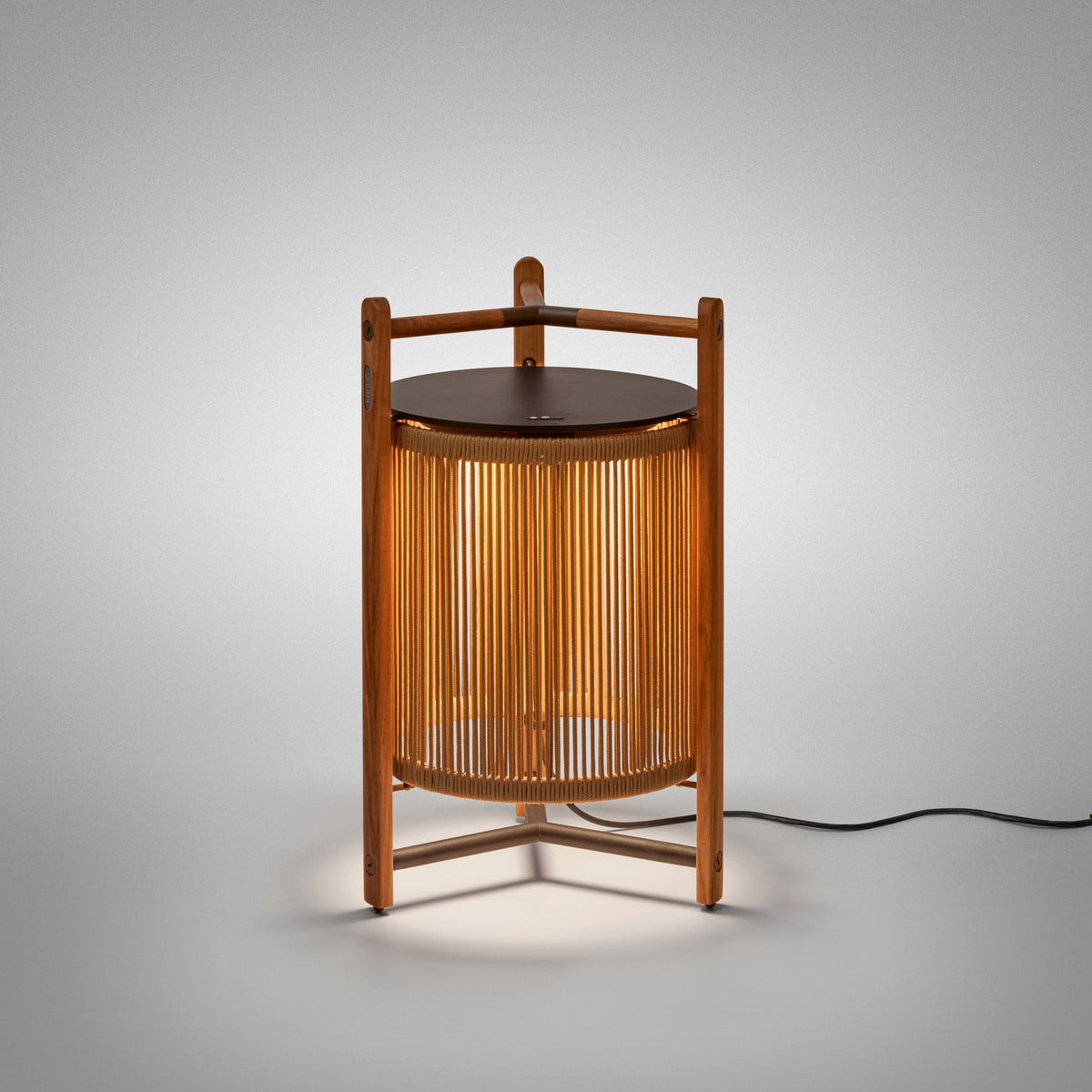 Kando P/76 outdoor floor lamp, lantern-shaped with sand nautical cord cylinder in a three-legged teak wood frame, dark round lid and wooden carry handle, lit on a plain grey background.