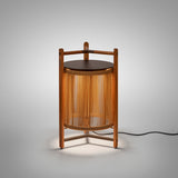 Kando P/76 outdoor floor lamp, lantern-shaped with sand nautical cord cylinder in a three-legged teak wood frame, dark round lid and wooden carry handle, lit on a plain grey background.