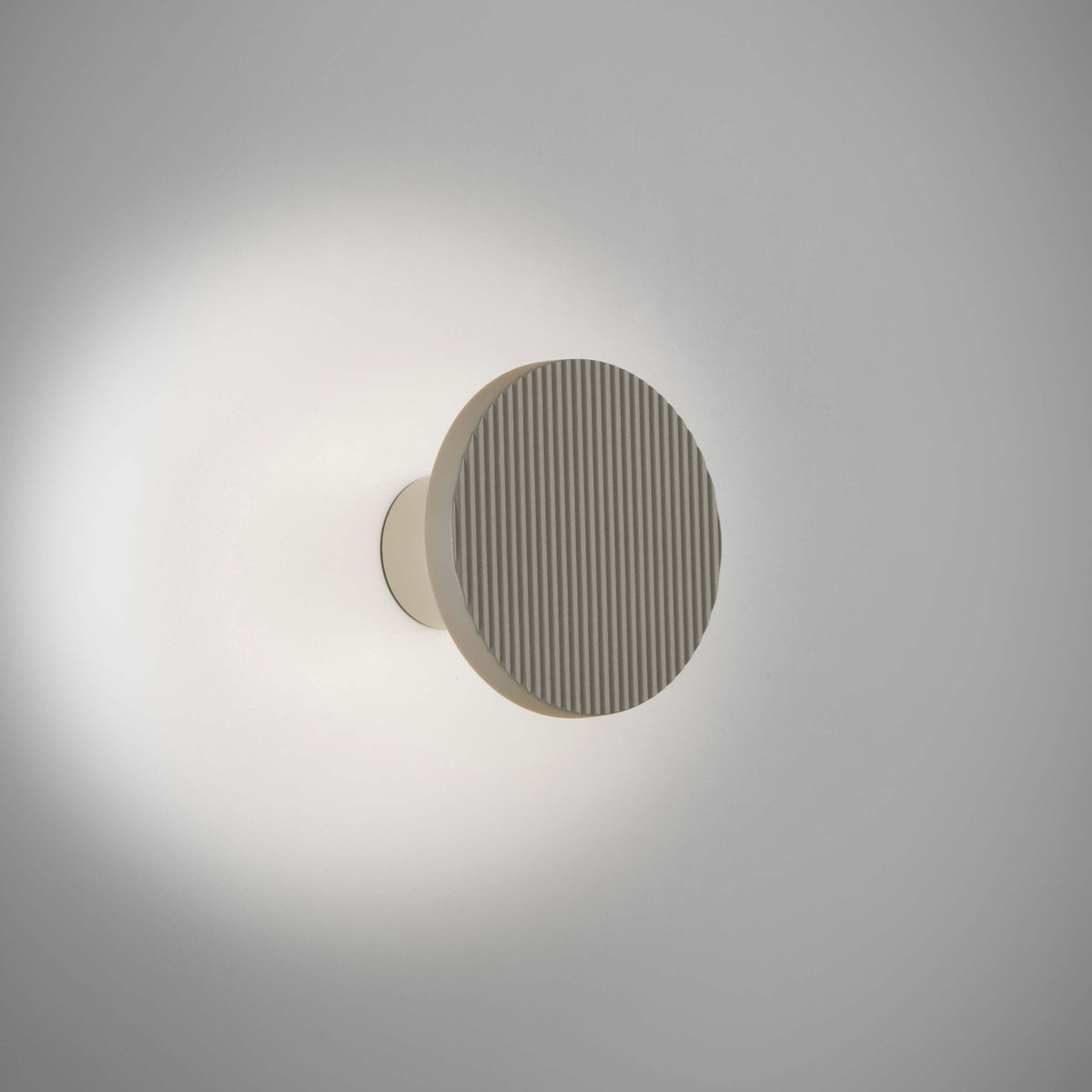 Arid A/01 round aluminium LED wall light with fine vertical fluting in Textured Pebble Grey, mounted on a short stem casting soft glow on a plain wall.