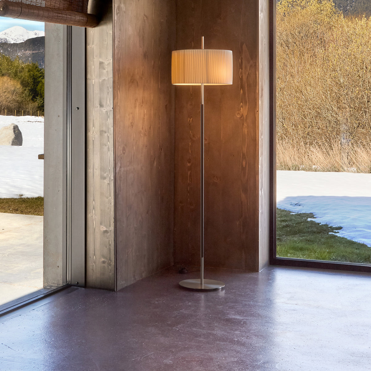 Danona P Fabric Drum Floor Lamp with pleated cream shade on a slim satin nickel stem and round base, shown lit in a wood-panelled room beside a window.