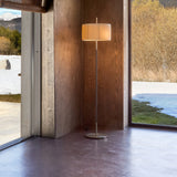 Danona P Fabric Drum Floor Lamp with pleated cream shade on a slim satin nickel stem and round base, shown lit in a wood-panelled room beside a window.