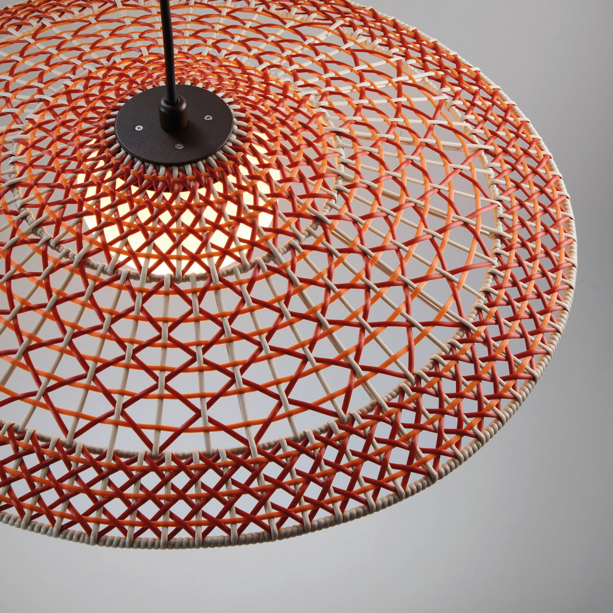 Close-up of the Nans S/55 woven LED outdoor pendant light with a wide shallow shade in red, orange, and beige fibre, graphite brown canopy, and warm white globe on a plain grey background.
