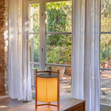 Kando M/40 Ribbon LED table lamp with cream pleated ribbon shade in teak wood frame, shown lit on a wooden table by curtained garden doors.
