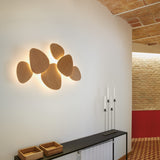 Tria 6 Light Wood LED Wall Light with pebble-shaped oak panels clustered on a white wall, backlit for a soft ambient glow, in a hallway setting.