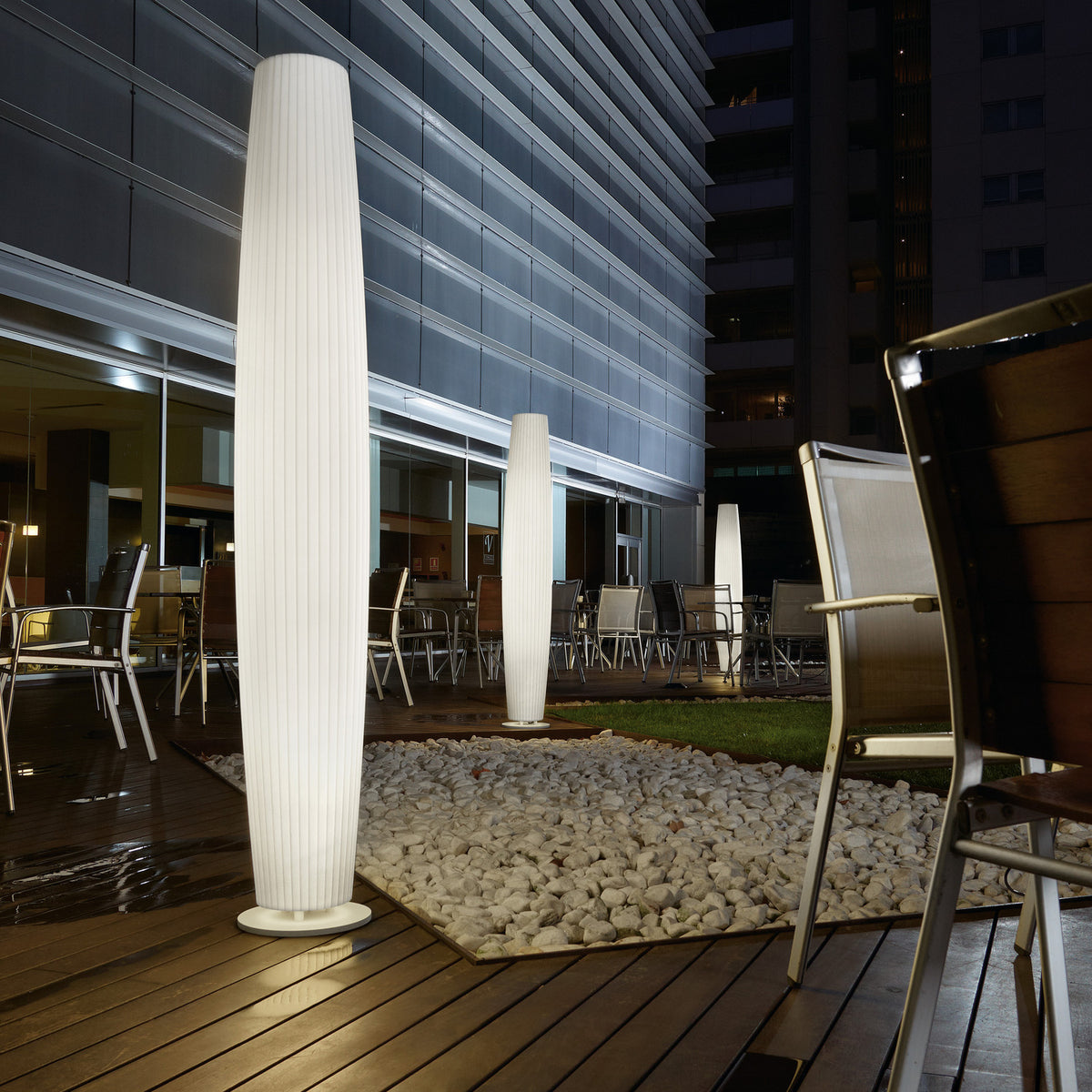 Maxi P/180 tall white polyethylene outdoor floor lamp with ribbed cylindrical shade and round base, illuminated on a restaurant terrace at night alongside a matching smaller model.