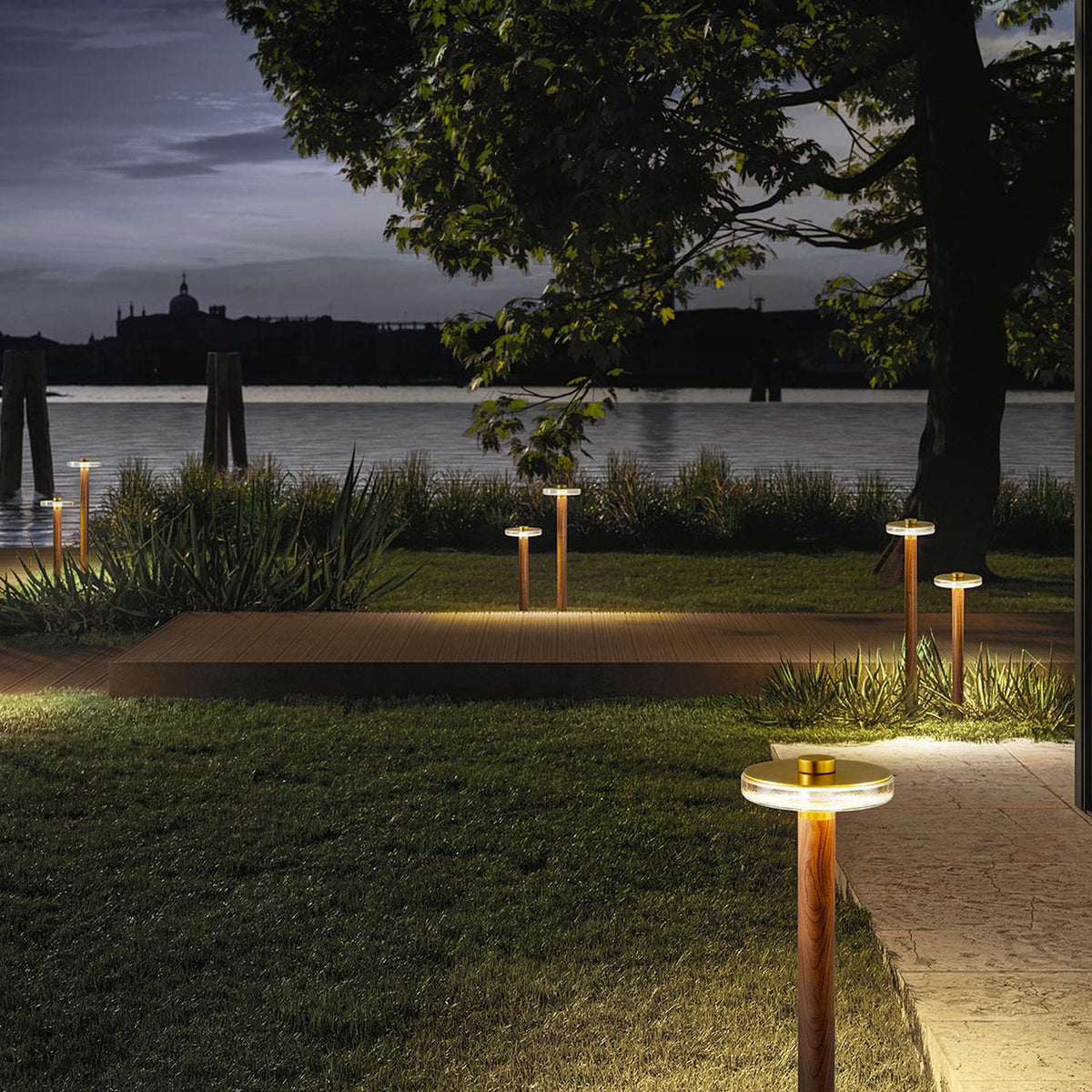 Venexia Glass LED Outdoor Floor Lamp 95cm in wood and matt brass, lit along a lawn and wooden deck beside a dusk waterside setting with a tree and distant skyline.