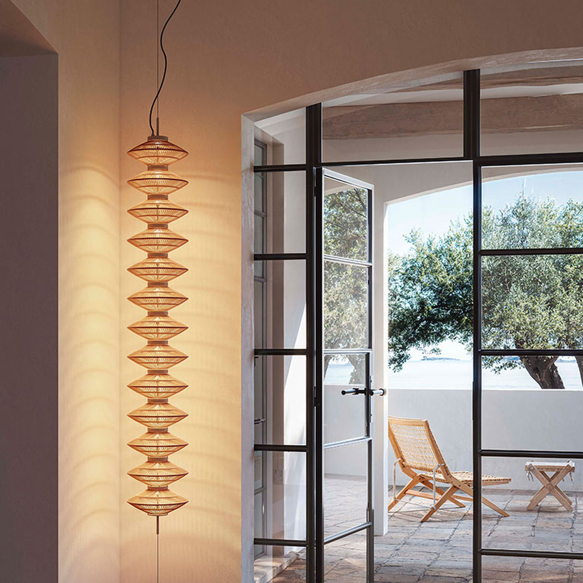 Yuyun S/11S fabric LED pendant light with eleven stacked woven beige cotton tiers on a slim stem, shown illuminated in a hallway beside glass doors opening to a terrace.