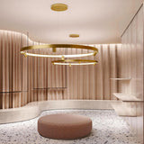 Arena Aluminium Ring LED Pendant Light 147cm in brass, two tiered circular rings with integrated LED band and spotlight heads, suspended in a warm-toned lounge setting.