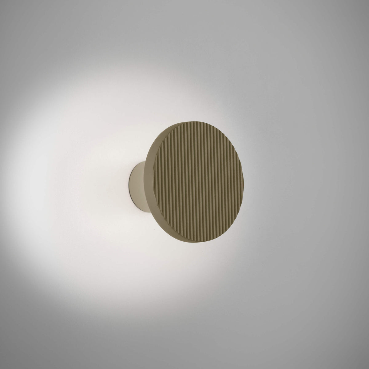 Arid A/01 round aluminium LED wall light with vertically fluted disc in Textured Olive Grey, mounted on a short stem casting a soft halo glow on a plain light background.