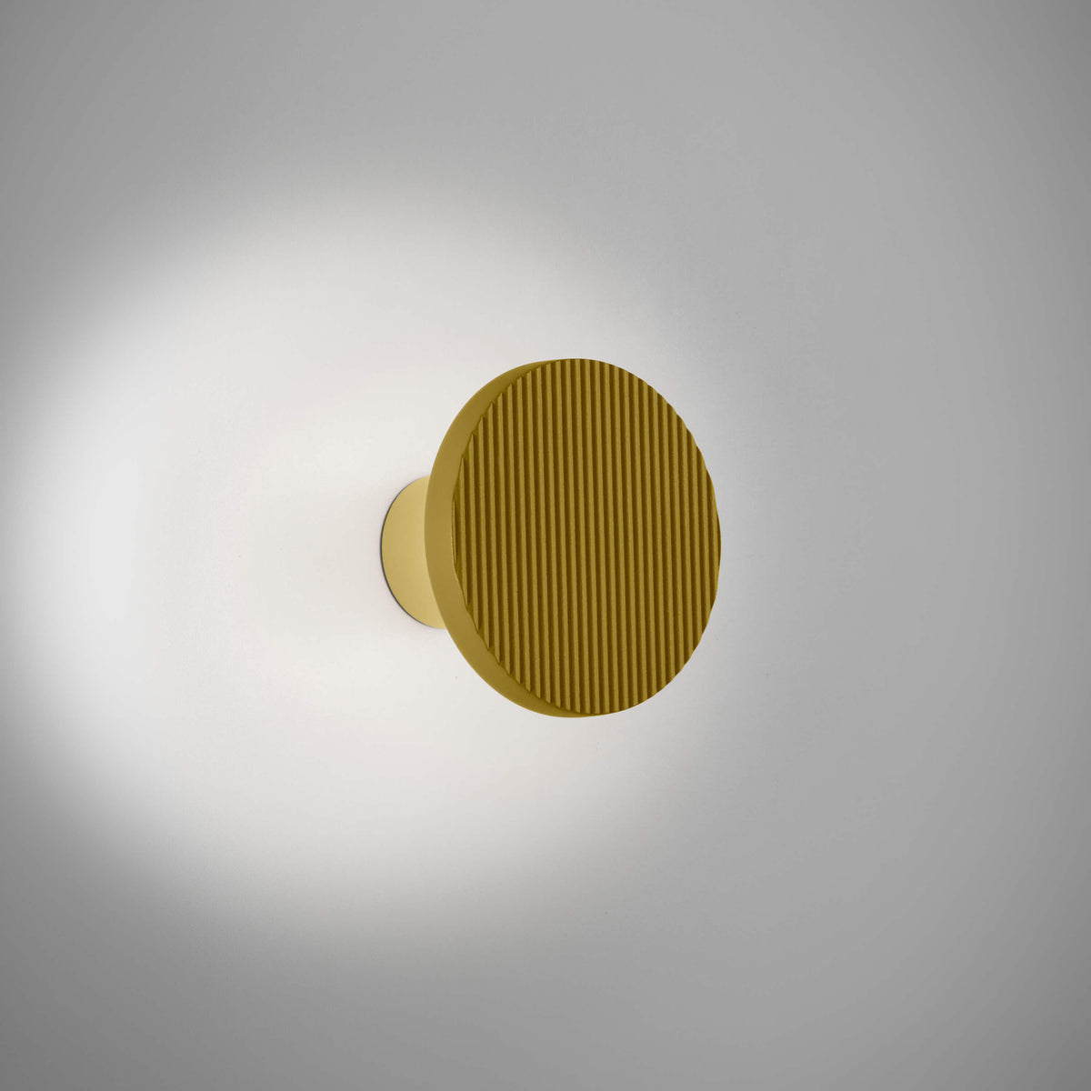 Arid A/01 round aluminium wall light with a ribbed sand yellow disc on a short stem, integrated LED casting a soft glow onto the wall, shown on a plain light background.