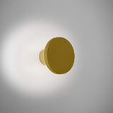Arid A/01 round aluminium wall light with a ribbed sand yellow disc on a short stem, integrated LED casting a soft glow onto the wall, shown on a plain light background.