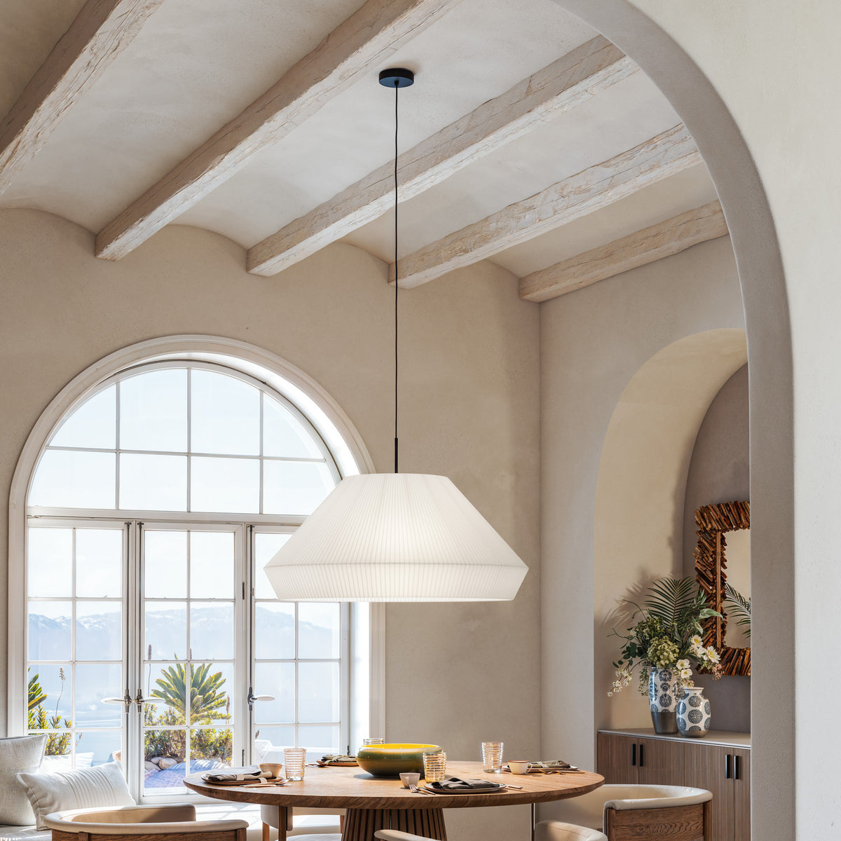 Mei 60 Ribbon Cone pendant light with a wide, pleated white translucent shade on a black cord, hung above a dining table beneath exposed ceiling beams.