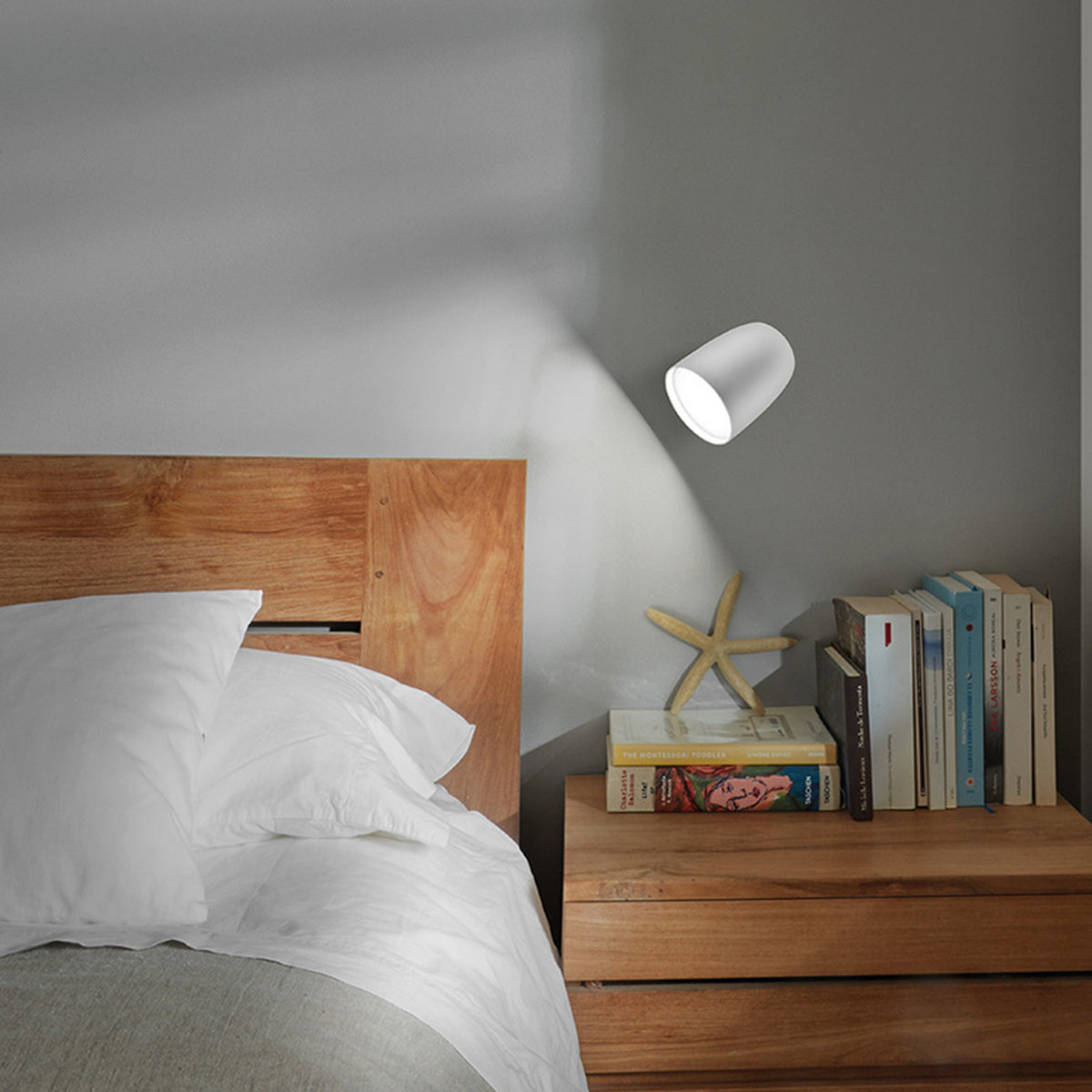Nut A/01 Stainless Steel LED Outdoor Wall Light in white, bell-shaped head on a slim wall plate, mounted above a wooden bedside table in a bedroom setting, casting warm light onto the grey wall.