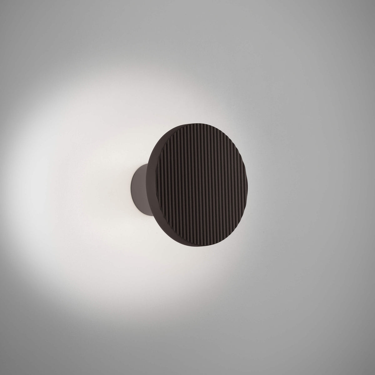 Arid A/01 round LED wall light in Textured Black aluminium, fluted disc on a short stem casting a soft halo of warm light onto a plain grey wall.